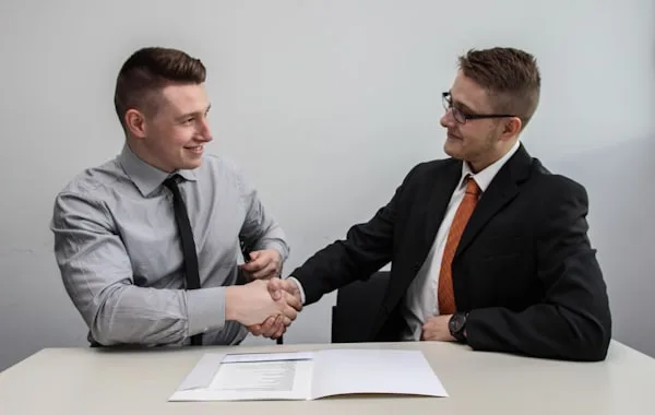 Two people shaking hands across a desk, agreement in front of them