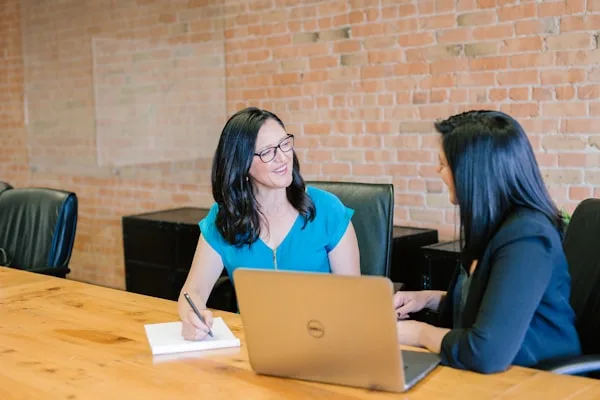 Two colleagues at a table, one talking the other through something on a laptop